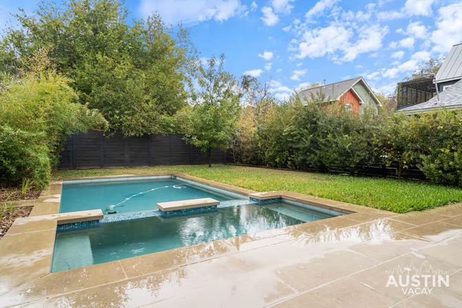 Residential backyard with a rectangular lap pool and attached spa, wet stone decking, green lawn and tall privacy hedges, nearby rooftops and a blue sky with scattered clouds.
