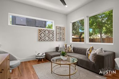 Bright Austin living room with gray L-shaped sofa, colorful patterned pillows, round glass-and-gold coffee table, wood floors, textured rug, modern wall art and large windows overlooking leafy trees.