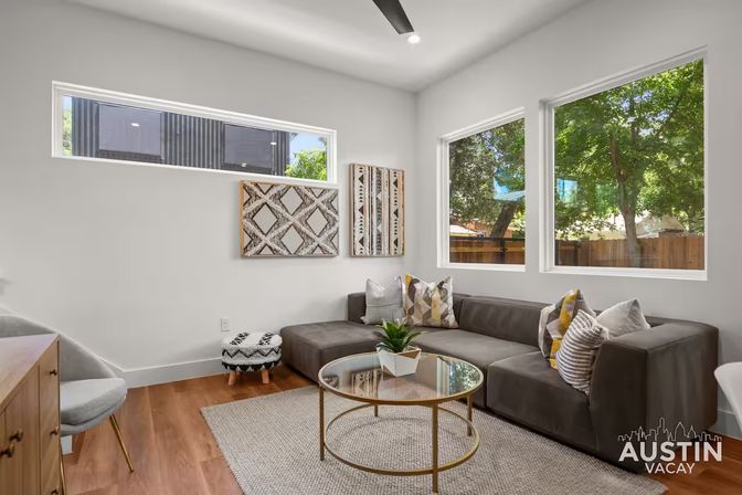 Bright Austin living room with gray L-shaped sofa, colorful patterned pillows, round glass-and-gold coffee table, wood floors, textured rug, modern wall art and large windows overlooking leafy trees.