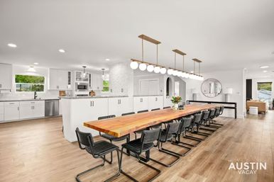 Bright open-plan Austin dining area with a long live-edge wooden communal table, black cantilever chairs, globe pendant lights, white kitchen cabinetry and warm hardwood floors, ideal for group gatherings.