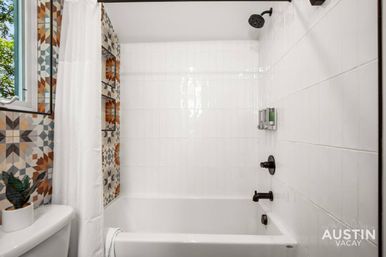 Bright bathroom with white tiled tub/shower, oil-rubbed bronze fixtures, colorful geometric accent tiles, wall dispensers, shower curtain, and potted plant by a sunlit window.