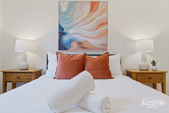 Cozy modern vacation rental bedroom with crisp white linens, two rust-colored throw pillows and rolled towels on the bed, wooden nightstands with ceramic lamps and an abstract painting above.