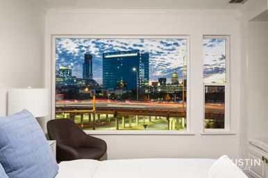Cozy bedroom window framing Austin skyline at twilight — glass towers and the capitol dome above a highway with streaking car light trails.
