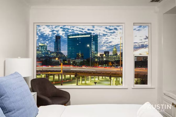 Cozy bedroom window framing Austin skyline at twilight — glass towers and the capitol dome above a highway with streaking car light trails.