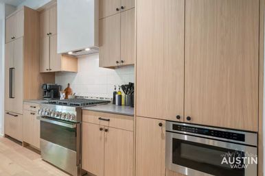 Modern light-wood kitchen in an Austin vacation rental with stainless steel gas range and built-in microwave, textured white backsplash, gray countertop with coffee maker and utensils.