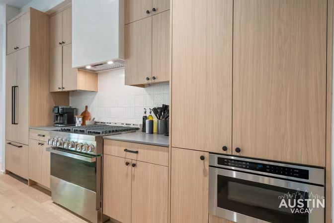 Modern light-wood kitchen in an Austin vacation rental with stainless steel gas range and built-in microwave, textured white backsplash, gray countertop with coffee maker and utensils.