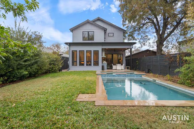 Backyard of a modern two-story gray home in Austin featuring a covered patio with outdoor seating, a rectangular swimming pool and spa, manicured lawn, and mature trees.