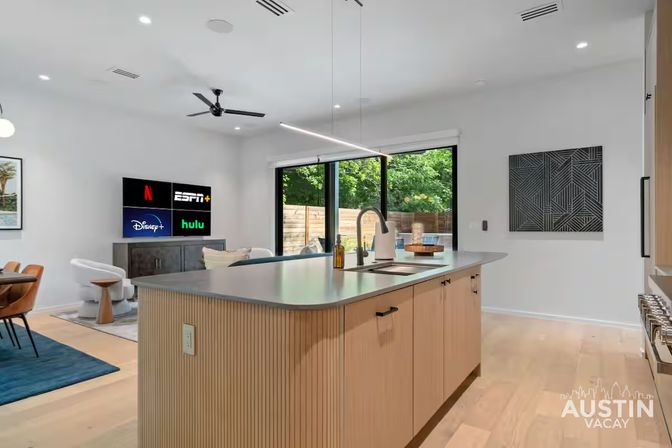 Bright modern Austin vacation-rental open-plan kitchen and living area with a light-wood island and sink, linear pendant light, sliding glass doors to a leafy fenced backyard, wall-mounted TV and dining nook.