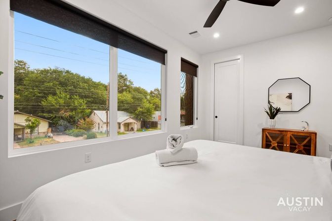 Bright modern Austin vacation rental bedroom with large windows overlooking a tree-lined neighborhood, crisp white bed topped with a rolled towel, wooden dresser with plant and octagonal mirror, and ceiling fan.