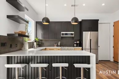Modern black-and-white kitchen with a large white island and four white bar stools, matte black cabinets, pendant lights, stainless steel appliances, hex tile backsplash and wood floors.