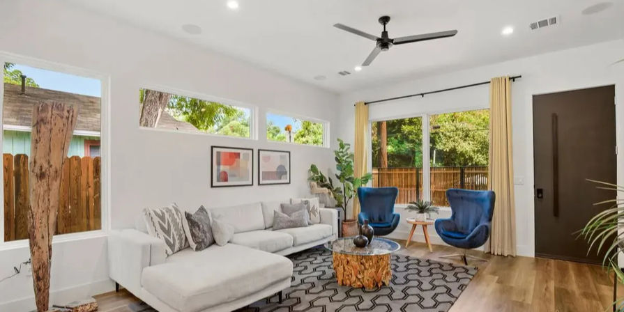 Bright contemporary living room with white sectional sofa, two blue accent chairs, wood stump coffee table, patterned rug, hardwood floors and large windows overlooking a leafy backyard.