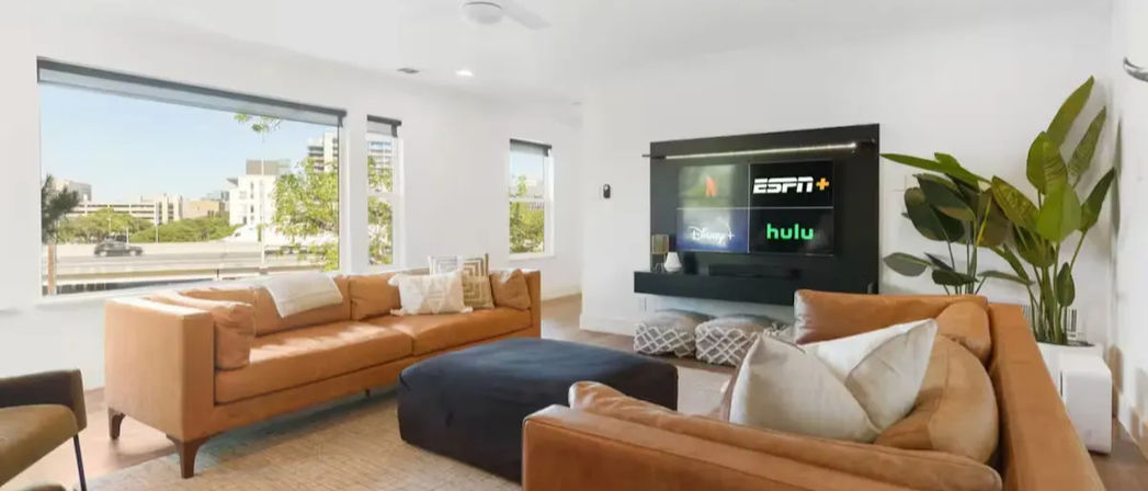 Cozy modern urban apartment living room with tan leather sofas, navy ottoman, large windows overlooking city buildings, wall-mounted TV showing streaming apps, indoor plant and neutral décor.