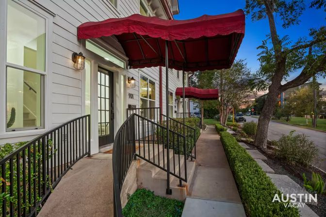 White townhome entrances with red canopy awnings, black metal railings and manicured hedges along a tree-lined city sidewalk at dusk in Austin