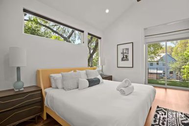 Sunlit modern bedroom with yellow upholstered headboard, crisp white bedding and decorative pillows, dark wood nightstands with ceramic lamps, high horizontal windows and sliding glass door to a balcony with leafy neighborhood views.