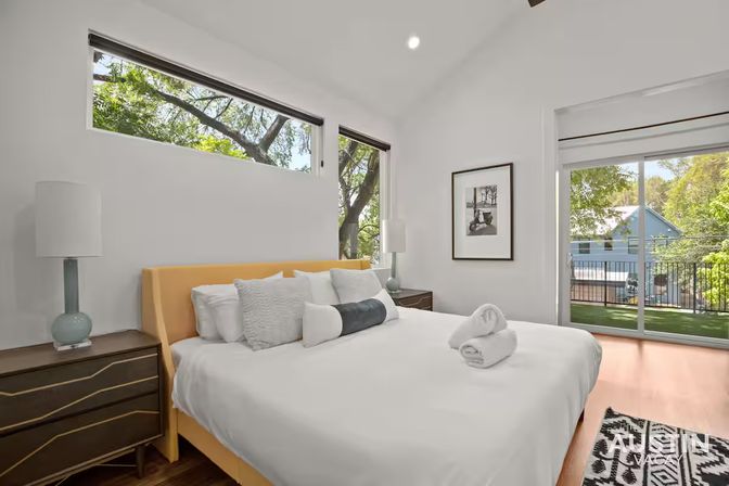 Sunlit modern bedroom with yellow upholstered headboard, crisp white bedding and decorative pillows, dark wood nightstands with ceramic lamps, high horizontal windows and sliding glass door to a balcony with leafy neighborhood views.