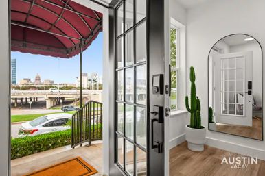 Open black-framed front door of a modern Austin apartment entry with a potted cactus, arched mirror, wood floors, orange doormat, and a downtown street view including the Texas State Capitol dome.