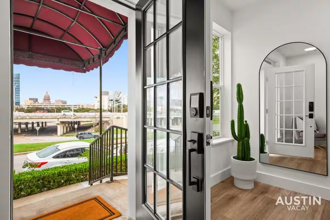 Open black-framed front door of a modern Austin apartment entry with a potted cactus, arched mirror, wood floors, orange doormat, and a downtown street view including the Texas State Capitol dome.