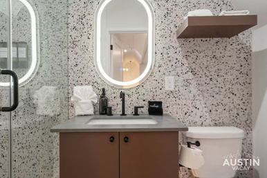 Stylish city apartment bathroom with terrazzo tile, backlit oval mirror, gray countertop over brown vanity, black faucet, glass shower door and white toilet.