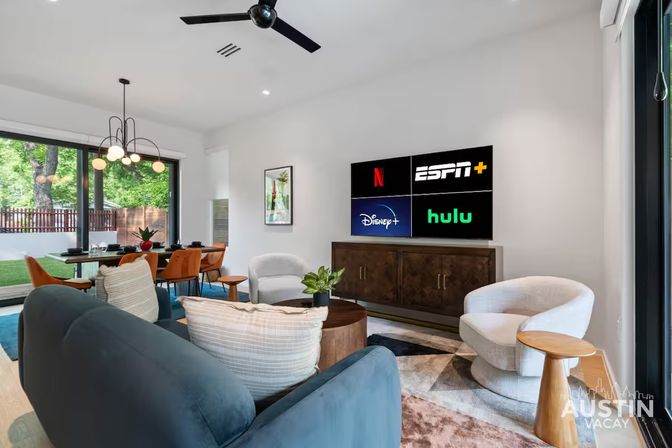 Cozy modern open-plan living room with blue sofa, white swivel chairs, dining table by large sliding glass doors overlooking a fenced backyard, and a wall-mounted TV showing Netflix, ESPN+, Disney+ and Hulu logos.