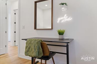 Cozy modern home office nook with black desk and mid-century wooden chair draped in a green throw, potted plant, wall mirror, and glowing 'good vibes' neon sign on a white wall over hardwood floors.
