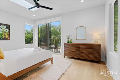 Bright modern Austin, Texas bedroom with wooden platform bed, sliding glass doors to a balcony and leafy street view, mid-century wooden dresser with mirror and lamp, skylight and black ceiling fan.