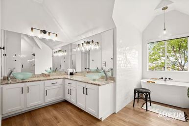 Bright modern bathroom with white L-shaped vanity and granite countertop, aqua glass vessel sinks under globe lights, large mirror, walk-in shower, freestanding tub by a window with tree view, and wood-look flooring.