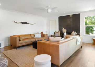 Contemporary sunlit living room with matching tan leather sofas, light wood floors, woven rug, black accent wall, ceiling fan, longhorn wall art and a large window with green view