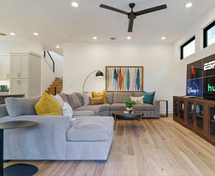 Bright modern open-plan living room with large gray sectional, colorful throw pillows, abstract wall art, round coffee table, hardwood floors and TV showing streaming apps