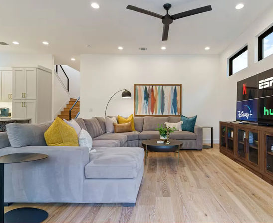 Bright modern open-plan living room with large gray sectional, colorful throw pillows, abstract wall art, round coffee table, hardwood floors and TV showing streaming apps