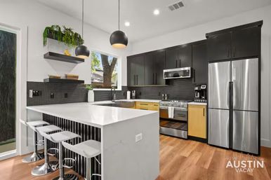 Bright modern vacation-rental kitchen with a white quartz island and three bar stools, matte black upper cabinets, light wood lower drawers, stainless steel fridge and range, round black pendant lights, small hex-tile backsplash and a window with a tree view.