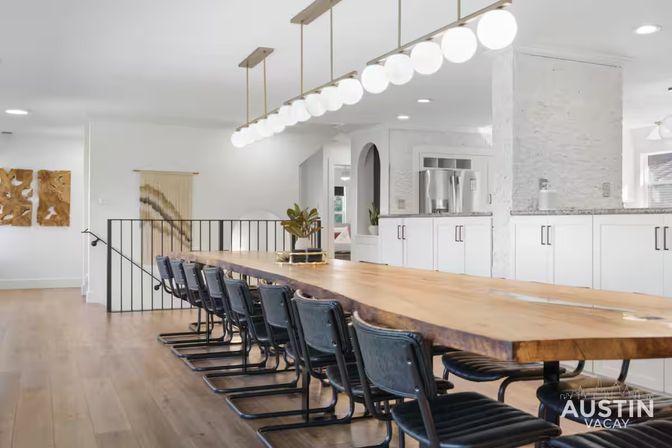 Spacious open-concept dining area with a long live-edge wooden communal table, black industrial chairs, globe pendant lighting, and a bright white modern kitchen in the background.