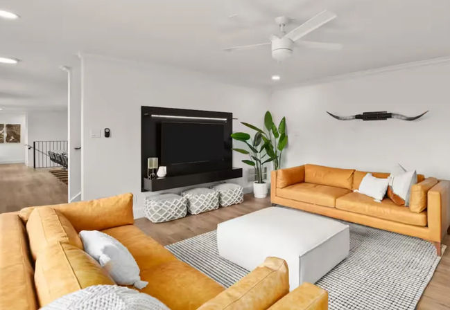 Bright modern living room interior with two mustard leather sofas facing a wall-mounted TV on a black media panel, white square ottoman on a gray patterned rug, potted tropical plant, decorative longhorn wall art, ceiling fan and light wood floors
