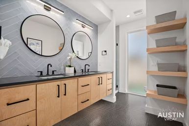 Modern bathroom with double vanity: light wood cabinets, black countertop and faucets, two round mirrors, gray herringbone tile backsplash, black penny-tile floor, open wood shelves with baskets