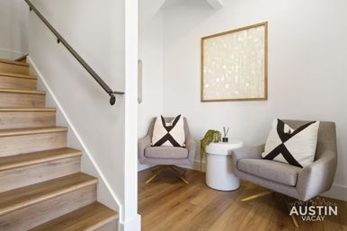 Inviting modern entryway with wood staircase and hardwood floors, two gray lounge chairs with black-and-white X pillows flanking a white round side table with a plant, neutral walls and framed abstract art.