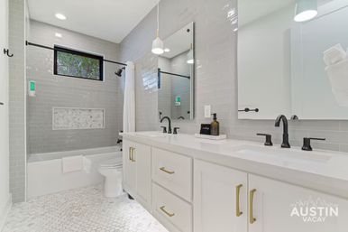 Bright modern bathroom with double white vanity and gold pulls, matte-black faucets, gray subway-tiled shower with marble niche, high window and hexagon marble floor.