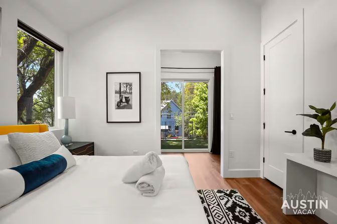 Bright modern bedroom in Austin — white walls, king bed with blue bolster and rolled towels, large window with trees and sliding glass door to backyard.