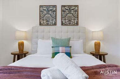Inviting modern bedroom with tufted upholstered headboard, colorful throw pillows and rolled white towels on crisp white bedding, wooden bedside tables with amber lamps and framed botanical prints above.