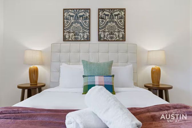 Inviting modern bedroom with tufted upholstered headboard, colorful throw pillows and rolled white towels on crisp white bedding, wooden bedside tables with amber lamps and framed botanical prints above.