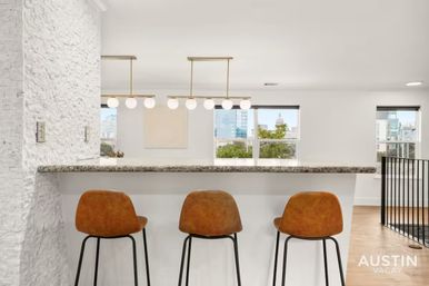Bright modern kitchen bar with granite countertop, three brown leather bar stools, globe pendant lights, and an urban skyline visible through wide windows.