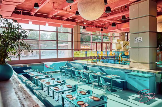 Vibrant indoor pool converted into a retro-style restaurant with turquoise tables and chairs set in a drained pool, pink ceiling, large windows and a potted plant, pool ladder and a 'No Diving' sign.