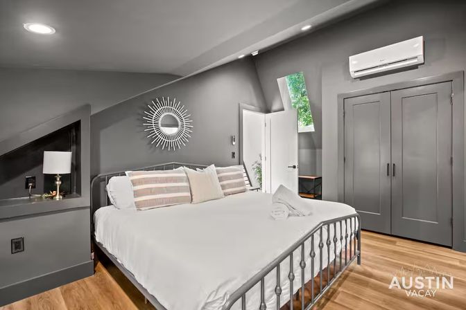 Stylish gray attic bedroom in an Austin vacation rental — metal-framed bed with white linens and striped pillows, sunburst mirror, hardwood floors, built-in lamp niche, wall-mounted AC and double closet