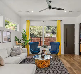 Bright modern living room with large window and yellow curtains, two blue accent chairs, white sofa, live-edge wood coffee table, geometric rug, hardwood floors, ceiling fan, and potted plant.