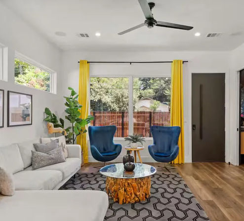Bright modern living room with large window and yellow curtains, two blue accent chairs, white sofa, live-edge wood coffee table, geometric rug, hardwood floors, ceiling fan, and potted plant.