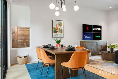 Modern Austin vacation-rental dining area with wooden table set for four, orange velvet chairs on a blue rug, globe chandelier, wall art and TV showing streaming app logos