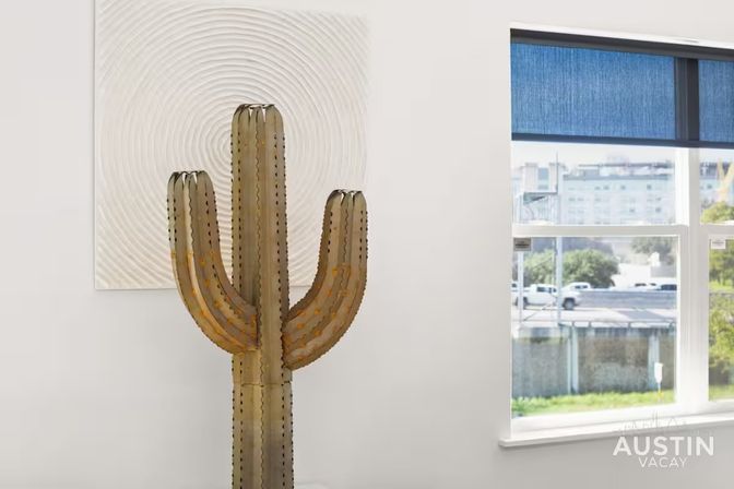 Metal saguaro cactus sculpture and textured circular wall art in a bright modern apartment, blue-shaded window revealing urban street and greenery outside