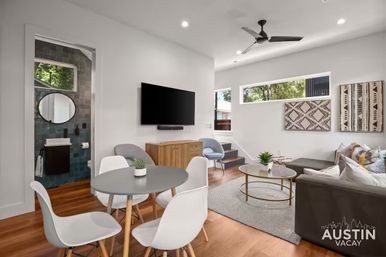 Bright Austin vacation-rental living room with hardwood floors, round gray dining table and white chairs, gray sectional sofa, wall-mounted TV, patterned wall art, gold-accent coffee table, ceiling fan, and a small tiled bathroom with round mirror visible through a doorway.