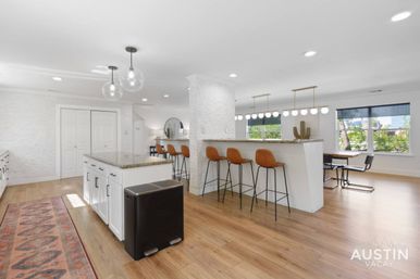 Bright, sunny open-concept kitchen and dining area with granite island, tan leather bar stools, globe pendant lights, hardwood floors, patterned rug runner, large windows and cactus decor.
