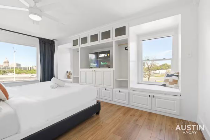 Sunlit modern bedroom in Austin with a king bed, white built‑in cabinetry and TV, cozy window seat, hardwood floors, and a city view including the Texas State Capitol dome