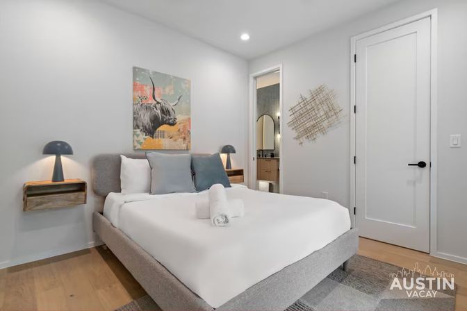 Bright modern bedroom with queen bed and crisp white linens, rolled towel on the bed, floating wooden nightstands with dome lamps, colorful highland cow artwork above the headboard, open doorway to an en-suite bathroom and light hardwood floors.