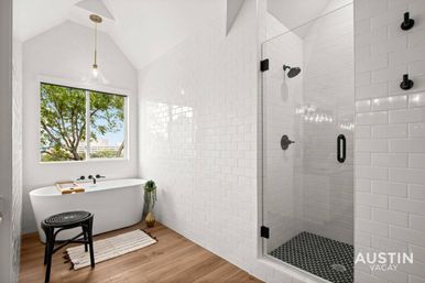 Bright modern bathroom with white subway tile, freestanding tub beneath a tree-view window, black matte fixtures, glass-enclosed shower with dark hex-tile floor, wood-look flooring, pendant light, stool and bath tray.